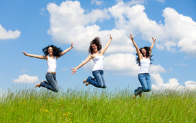 Три молодые девушки прыгают в зеленой траве на фоне неба 