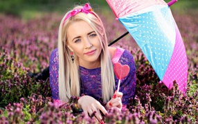 Молодая девушка блондинка лежит на поляне с сиреневыми цветами под разноцветным зонтом