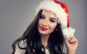 Beautiful long-haired brunette girl in a Santa Claus hat on a gray background