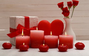 Зажженные красные свечи на столе с подарком, красным сердцем и цветами в вазе 