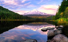 Заснеженная гора отражается в воде озера у зеленого леса