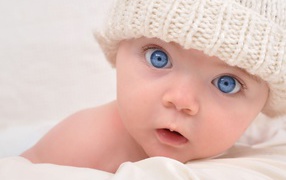Голубоглазый малыш в белой вязаной шапке на сером фоне