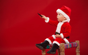Little boy dressed as Santa Claus sits on a chair on a red background