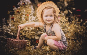 Little girl in a hat sits on the grass with a basket