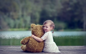 Маленькая девочка в белом платье сидит на мосту с плюшевым медведем