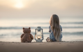 Маленькая девочка сидит на песке с плюшевым мишкой 