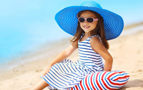 Маленькая девочка сидит на подушке на пляже в большой синей шляпе
