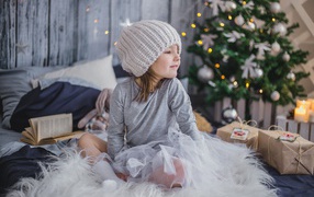 Маленькая девочка сидит на кровати у новогодней елки