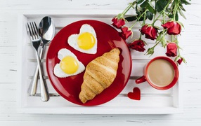 Fried eggs with croissant on a red plate on the table with a bouquet of roses and a cup of cocoa