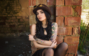 Девушка в шляпе с татуировками на руке сидит у стены