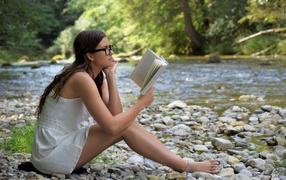 Молодая девушка в белом платье читает книгу на берегу реки