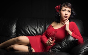 Девушка в красном платье с бокалом в руке лежит на кожаном диване