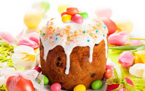 Красивый пасхальный кулич с белой глазурью и конфетами на столе с яйцами