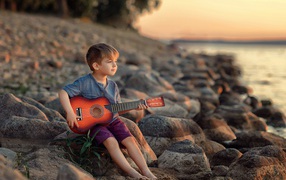 Маленький мальчик сидит с гитарой на камнях у моря 