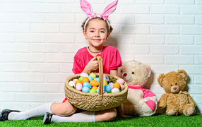 Улыбающаяся девочка сидит у стены с корзиной пасхальных яиц и игрушками