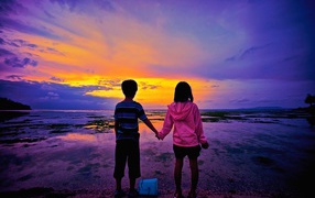 Маленькие мальчик и девочка стоят у воды на фоне заката 