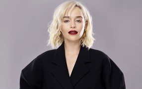 Яркая блондинка, актриса Эмилия Кларк в черном пальто на сером фоне