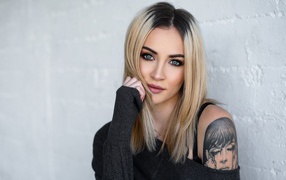 Стильная блондинка с татуировкой на плече на фоне стены