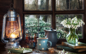 Керосиновая лампа на столе с кружкой, книгами и букетом подснежников у окна