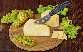 A piece of hard cheese on a table with a knife and white grapes