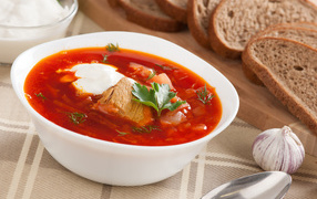 A plate of hot borsch on a table with bread and garlic