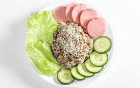 Гречка на тарелке с огурцом, колбасой и листьями салата на белом фоне