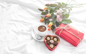 Cup of coffee on a white bed with sweets, a gift and a bouquet of roses