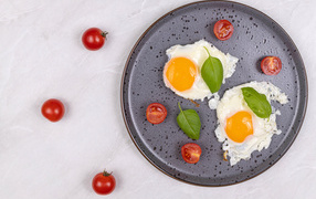 Fried eggs with tomatoes and basil leaves on a black plate