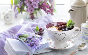 Muesli with berries in a cup on a table with a notebook and a bouquet of lilacs