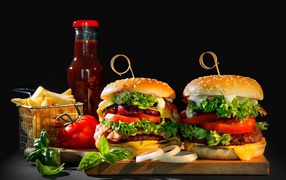 Two hamburgers on the table with french fries, ketchup and tomatoes