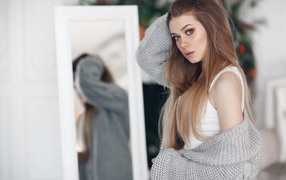 A young girl in a gray sweater stands at the mirror