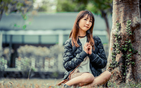 Девушка азиатка в черной куртке сидит под деревом