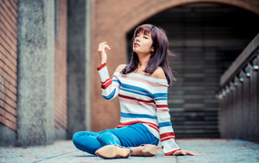 Девушка азиатка в джинсах и полосатом свитере сидит на асфальте