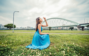 Красивая девушка азиатка в голубом платье  сидит на траве на фоне моста