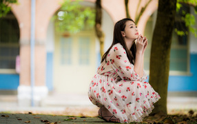 Красивая девушка азиатка в платье сидит под деревом в парке