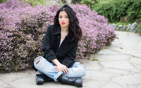 Красивая девушка в черном пальто сидит на земле у кустов