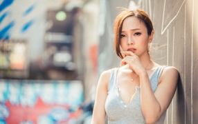 Милая девушка азиатка  с рукой у лица стоит у стены 