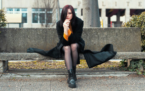 Девушка в черном пальто сидит на бетонной лавке 