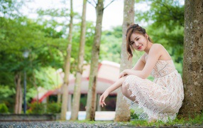 Улыбающаяся девушка в кружевном платье сидит под деревом в парке