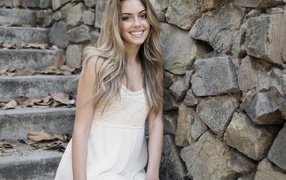 Улыбающаяся  девушка в белом платье стоит на ступеньках у стены