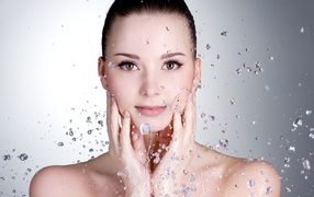 Брызги воды летят на красивое лицо молодой девушки на сером фоне