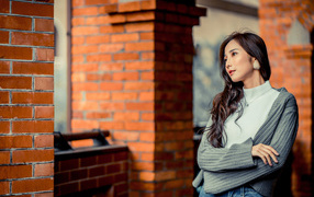 Молодая девушка азиатка в серой кофте у кирпичной стены 