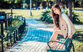 Молодая девушка азиатка сидит в пальто на лавке в парке 