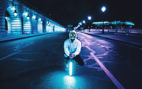 A guy in a luminous mask sits with a neon lamp on the road