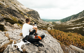 Парень с квадрокоптером сидит на камне в горах 