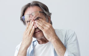 Пожилой мужчина трет глаза на сером фоне