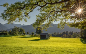 The barn stands on a green meadow in the sun against the backdrop of mountains