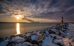 Snow-covered stones by the sea with a lighthouse at sunset