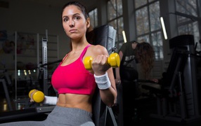 Sports girl shakes her hands with yellow dumbbells in the gym