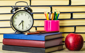 Будильник, книги, карандаши и яблоко на столе у стены 
